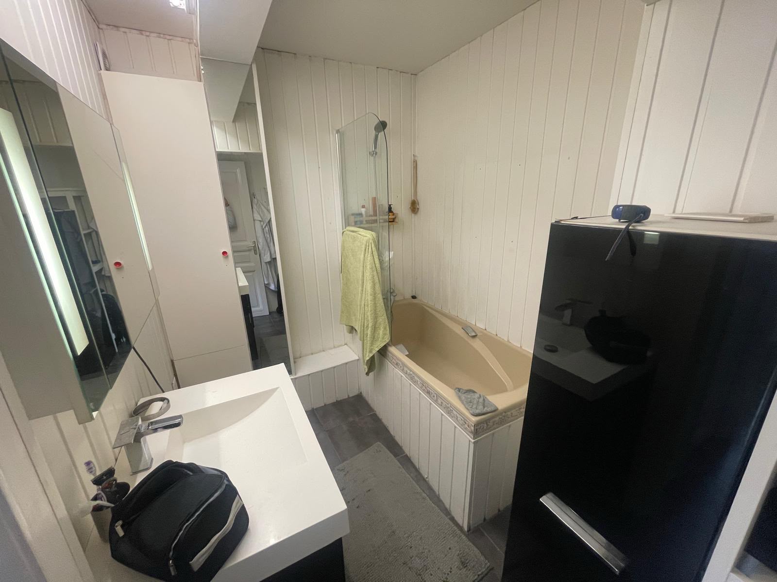 Avant rénovation : vue d’ensemble de la salle de bains avec meuble vasque blanc, baignoire beige et parement mural blanc.