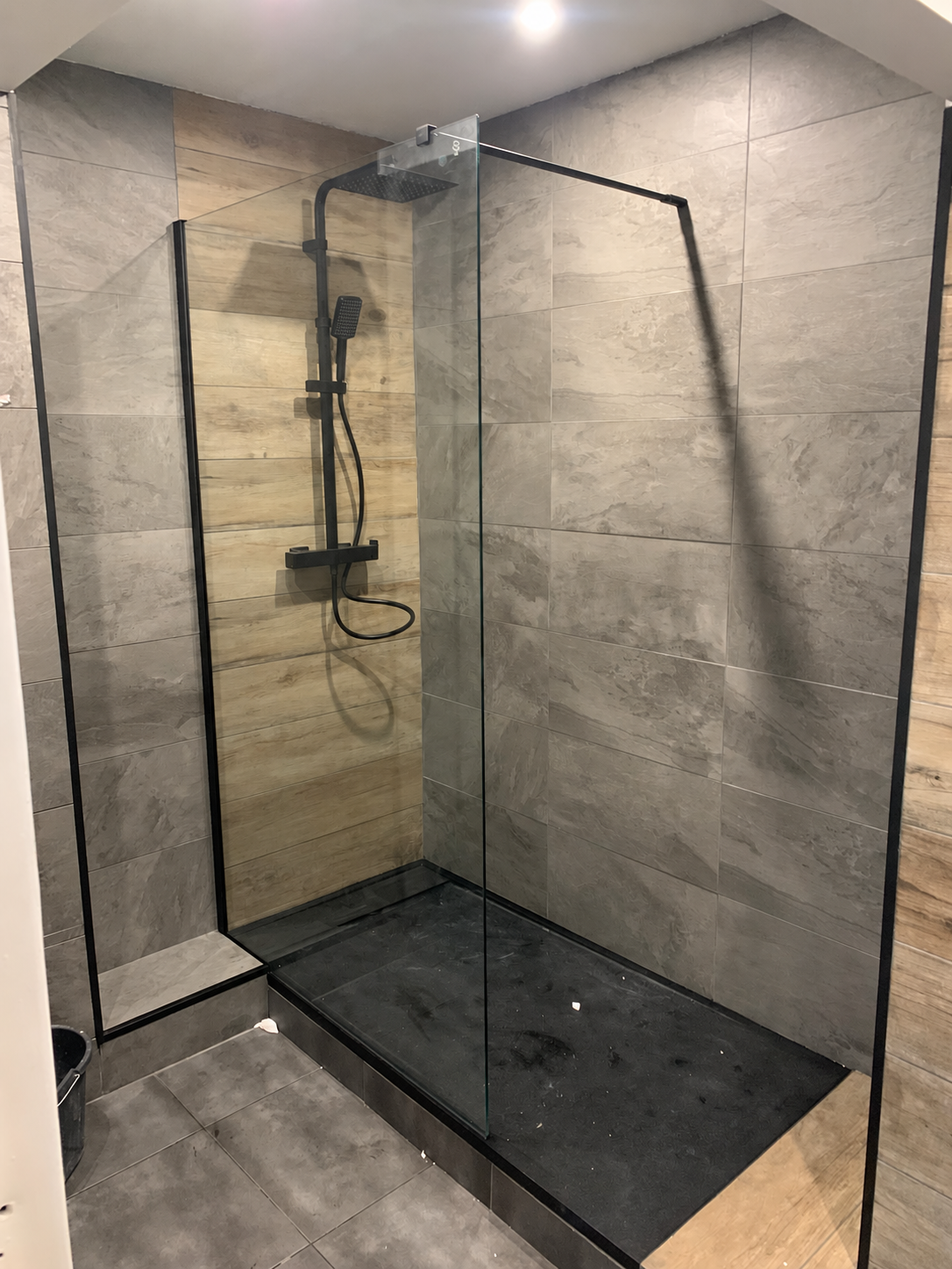 Après rénovation : douche avec receveur et robinetterie noirs, paroi vitrée, carrelage gris et finitions effet bois.
