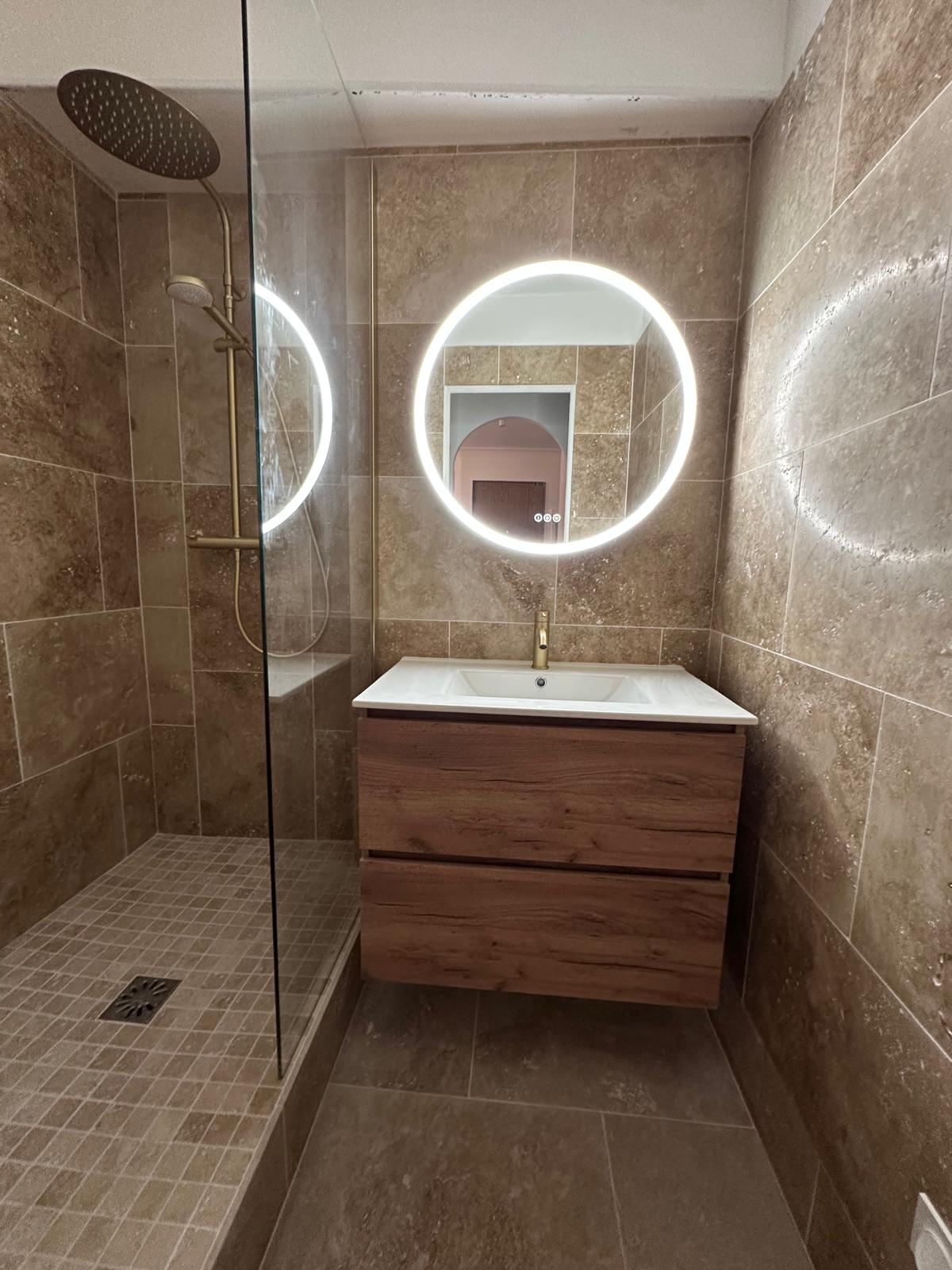 Après rénovation : douche vitrée, meuble vasque en bois et miroir rond lumineux.